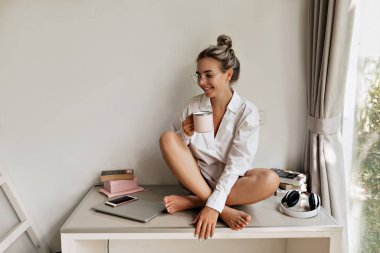 Masada dizüstü bilgisayarlı, akıllı telefonlu, beyaz tişörtlü ve gözlüklü, modern stüdyoda bir fincan kahveyle kitap okuyan hoş bir kadın. 