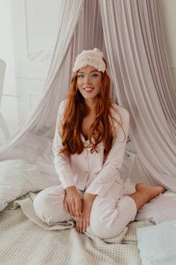 Çekici kızıl saçlı, uzun dalgalı saçlı, pembe pijamalı bir kadın. Sabahları yatağında güneş ışığında oturuyor.