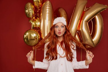 Uzun dalgalı saçlı, çekici, parlak bir kadın kırmızı arka planda kameraya bir öpücük yolluyor. Fotoğrafta pembe elbiseli kadın kameraya bakıyor.