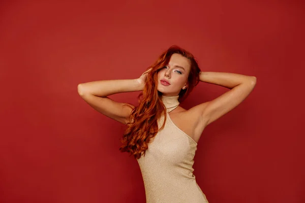 Elegante mujer elegante en vestido de oro posando sobre fondo rojo tocando su cabeza —  Fotos de Stock