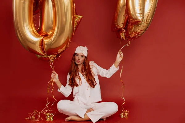 Studio portrait of brightful woman posing with golden balloons over red background wearing pink sleeping suit and mask — Stock Photo, Image