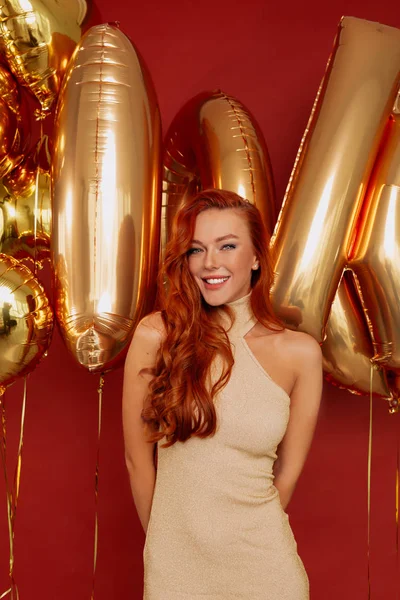 Sorridente ragazza dai capelli rossi felice indossa abito da sera in oro posa su sfondo rosso con palloncini d'oro . — Foto Stock