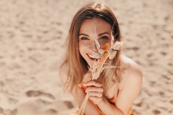 Close up fora retrato de encantadora linda senhora cobrindo rosto com flores de verão e sentado na praia de areia sob a luz do sol — Fotografia de Stock
