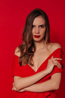 Confident pretty woman with wavy hair looking at camera wearing red dress with red lips over red background. Stylish womans portrait 