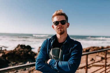 Güneş gözlüklü yakışıklı genç adam güneşli bir günde mavi gökyüzüne ve okyanus dalgalarına karşı poz veriyor. Deniz kenarında yürüyen ciddi bir genç adam..