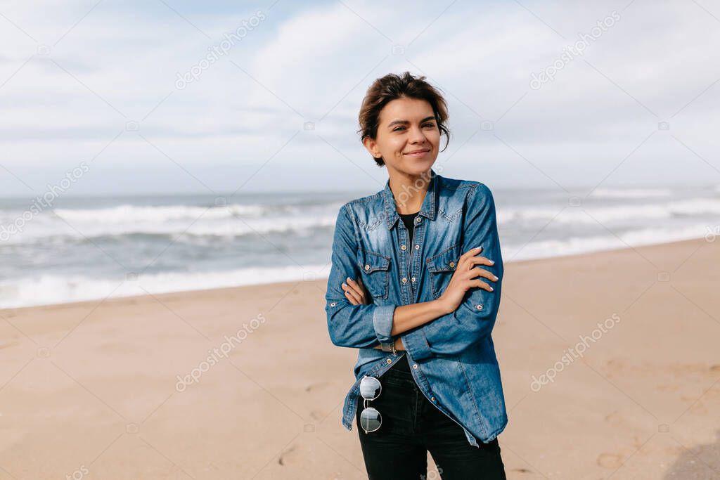 Young attractive smiling woman in denim shirt walking by the ocean against blue sky in sunny day. 