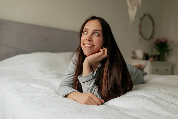 Alegre chica feliz relajándose en la cama blanca por la mañana. Joven alegre con el pelo largo morena soñando y sonriendo —  Fotos de Stock