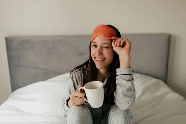Tutup potret wanita menawan tersenyum di tempat tidur dengan secangkir kopi mengedipkan mata pagi dan memegang topeng tidur — Stok Foto
