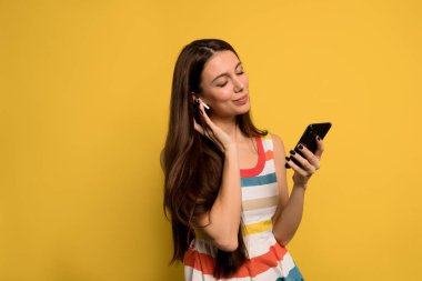 Uzun siyah saçlı, modern, hoş bir kadın. Parlak bir elbise giyiyor. Sarı duvarda akıllı telefonlu müzik dinliyor.. 