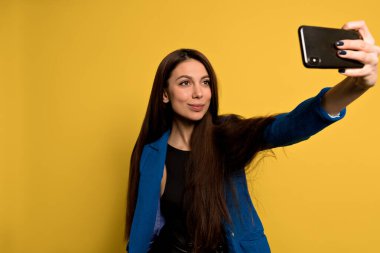 Uzun siyah saçlı, şık modern kadın mavi ceketli, izole edilmiş sarı arka planda akıllı telefonuyla selfie çekiyor.. 