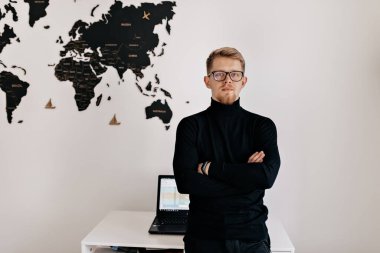 Gözlüklü, siyah kazaklı yakışıklı bir adamın dünya haritası ve dizüstü bilgisayarla beyaz duvarın üzerinde poz verirken çekilmiş kapalı bir portresi.. 