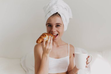 Kafasında havluyla mutlu, çekici genç bir kadın. Beyaz bluzlu, ısıran kruvasan giymiş. Yatakta izole edilmiş arka planda dinleniyor.. 