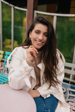 Uzun dalgalı saçlı, beyaz ceketli, açık terasta oturan ve arkadaşlarını bekleyen hoş bir kız. Mutlu bir kadının dış portresi . 