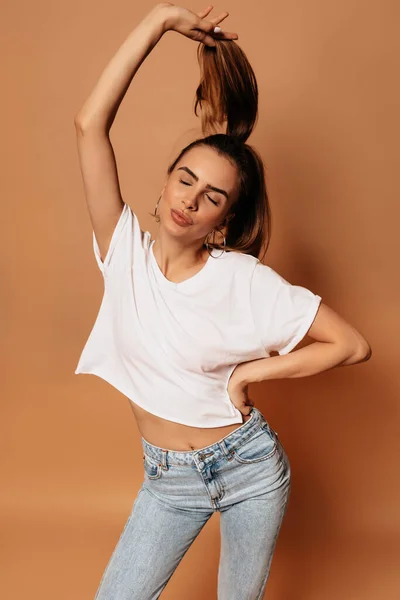 Studio colpo di attraente donna elegante indossare bianco t-shirt e jeans divertirsi e fare volti su sfondo beige — Foto Stock