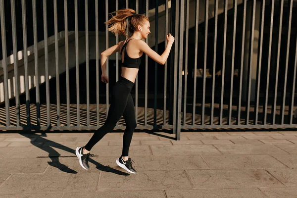 Ganzes Porträt einer athletischen Sportlerin in Sportuniform, die im Sonnenlicht mit Kopfhörern läuft. Sommersport und Freiheitskonzept. Athletentraining an sonnigem Tag — Stockfoto