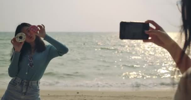 人生を楽しむ女性の友人は ビーチでリラックスして自由に写真を撮る甘いおいしいドーナツを食べて アジアの女性旅行者のライフスタイルの魅力的な一緒に Uhdフッテージ映画 — ストック動画