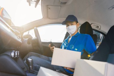 Maske takan Asyalı Teslimatçı, alıcının önünde akıllı telefon taşıyan bir paket gönderdi. Virüs tıbbi konsepti yayarken, bir sürü paket hatchback vagonunda kargo sosyal mesafesini teslim etti..