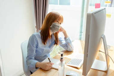 Maske takan Asyalı bir kadın son teslim tarihi olan karantina çalışma masasını bilgisayar ve koruyucu dezenfektanla strese soktu, el jeli alkolü temizledi, koronavirüsün yayılmasını engelledi..