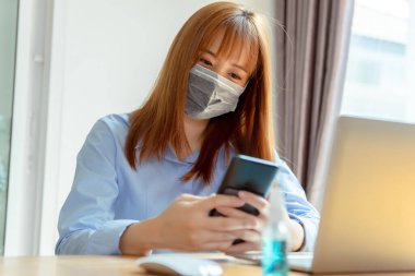 Maske takan Asyalı kadın evde kalıp akıllı telefonları dinlendiriyor, Coronavirus karantina çalışma masasının bilgisayar ve koruyucu maskeyle yayılmasını engelliyor..
