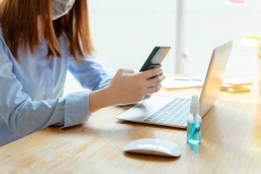 Maske takan Asyalı kadın evde kalıp akıllı telefonları dinlendiriyor, Coronavirus karantina çalışma masasının bilgisayar ve koruyucu maskeyle yayılmasını engelliyor..