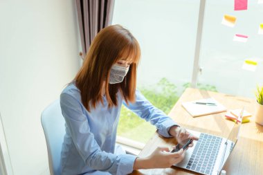 Maske takan Asyalı kadın evde kalıp akıllı telefonları dinlendiriyor, Coronavirus karantina çalışma masasının bilgisayar ve koruyucu maskeyle yayılmasını engelliyor..