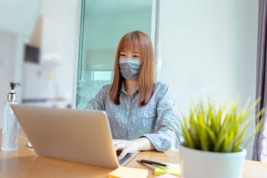 Maske takan mutlu bir Asyalı kadın evde online eğitimde çalışıyor, Coronavirus karantinasının covid-19 için yayılmasını engelliyor, riskleri azaltmak için toplumdan kendini izole ediyor..