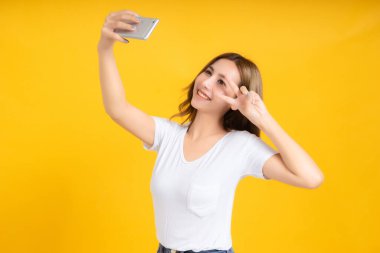 Portre mutlu genç Asyalı kadın gülen parmak el işareti göz jesti selfie eğlenceli komik pozitif duygu beyaz tişört, sarı arka plan izole stüdyo çekimi ve uzay kopyası.