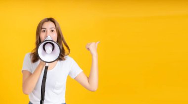 Yüksek sesle konuşan, Asyalı genç bir kadının beyaz tişörtlü, megafon reklamlı, sarı arka plandaki, izole edilmiş stüdyo fotoğraflarında gülümseyen,.