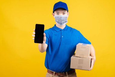 Çevrimiçi alışveriş için cep telefonlu maskeli yakışıklı bir adam. Paket, ev alıcısına teslim edilecek. Sarı arka plandaki izole edilmiş stüdyoda virüs bulaşmasını önleyen kargo teslimatı..