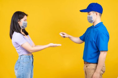 Maske takan Asyalı Teslimatçı, virüs sarı arka planda yayılıyorken kargo sosyal mesafesini dağıtmadan önce müşteriye el spreyi ile alkol temizliği uygulayan bir paket gönder..