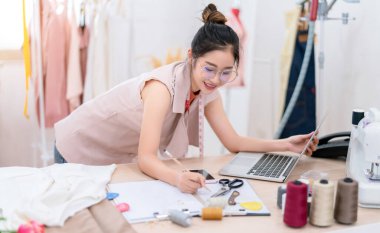 Güzel Asyalı kadın moda tasarımcısı kumaşı taslak kıyafetlerini dizüstü bilgisayarla müşteriye dikiyor, stüdyo terzisinde yaratıcı, meslek sahibi küçük işletme sahibi. 