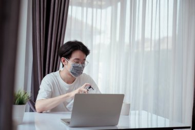 Maske takan Asyalı adam el pompası jeli temizlik dezenfektanı evde dizüstü bilgisayarla çalışıyor. Koronavirüs karantinasının yayılmasını engelliyor..