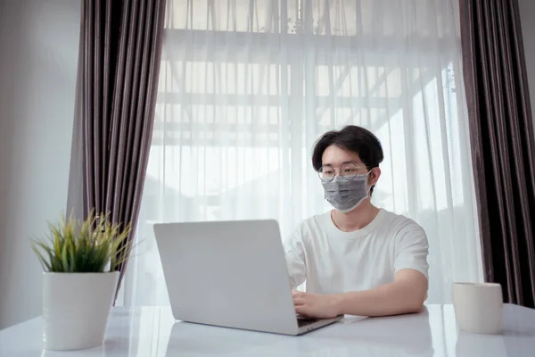 Asian man wearing mask working from home with laptop computer self isolation from society to reduce risk, quarantine prevention coronavirus, covid-19 and study e-learing online education technology.