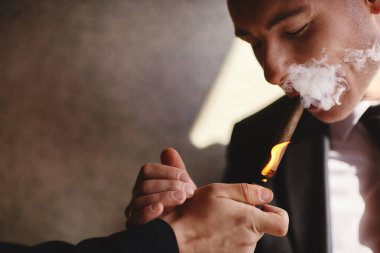 Takım elbiseli ve beyaz gömlekli şık bir adam kapalı mekanda puro içiyor, yakın plan, seçici odaklanma. Puro dumanı