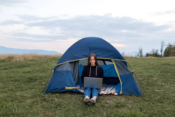 Girl with laptop in the mountains. Tourist woman uses a laptop in nature, freelancer typing on a computer near a tent in a journey against the backdrop of a mountain landscape — Stock Photo, Image