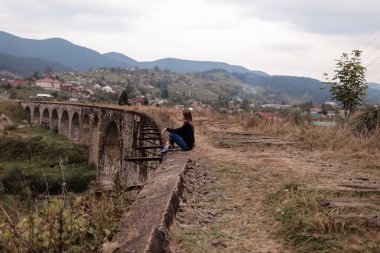 Eski tren raylarıyla viyadük üzerinde oturan genç turist kız. Vorokhta dağ beldesi köyünde eski bir viyadük. Ukrayna, Karpatlar.