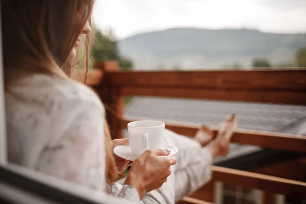 Junge Frau in stylischer Nachtwäsche trinkt morgens gerne Kaffee oder Tee im Freien auf dem Balkon und blickt in die Berge. — Stockfoto