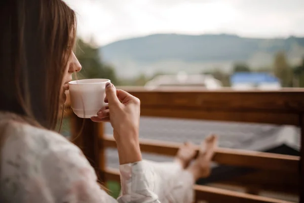 Junge Frau in stylischer Nachtwäsche trinkt morgens gerne Kaffee oder Tee im Freien auf dem Balkon und blickt in die Berge. — Stockfoto