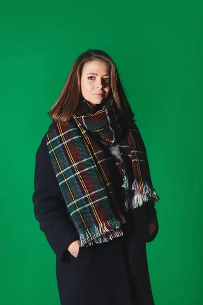 Молода красива дівчина в чорному пальто і шарф в клітці. Симпатична модель в теплому зимовому одязі позує на зеленому тлі. сезон зимових канікул . — стокове фото
