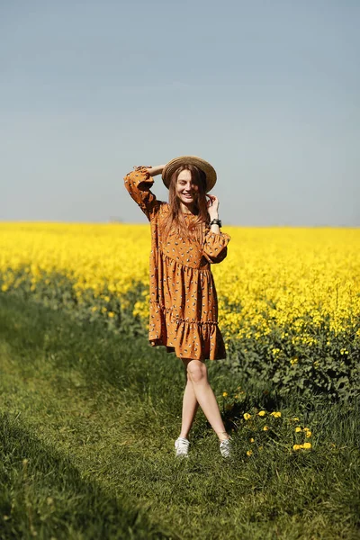 Stilren ung kvinna i halmhatt i ett fält av gula blommor. Flicka i blommig klänning. bakgrund med gula blommor och blå himmel — Stockfoto