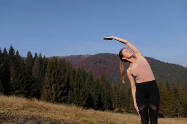 Dağlarda doğa üzerinde esneme egzersizleri yapan genç bir kadın. Spor kızı taytla yoga pozu veriyor. Güzel orman manzarası