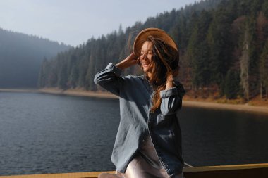 Şapkalı mutlu genç kadın ormandaki göl manzarasının tadını çıkarıyor. Rahatlatıcı anlar. Havalı kız manzarası açık havanın tadını çıkarıyor. Özgürlük, insanlar, yaşam tarzı, seyahat ve tatil