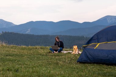 Genç bayan serbest yazar akşamları dağlarda dizüstü bilgisayarda çalışıyor. Kamp ateşi ve çadırın yanında oturan turist kız. Boşluğu kopyala.