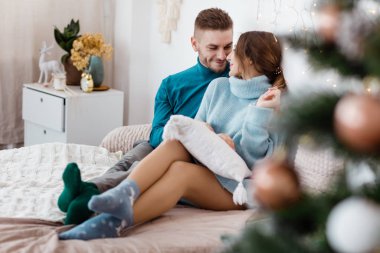 Evde Noel süslemesi yapan mutlu bir çift. Yeni yıl arifesi, süslenmiş köknar ağacı. Kış tatili ve aşk konsepti. Genç mutlu çift sarılıyor ve rahat koltukta dinleniyor..