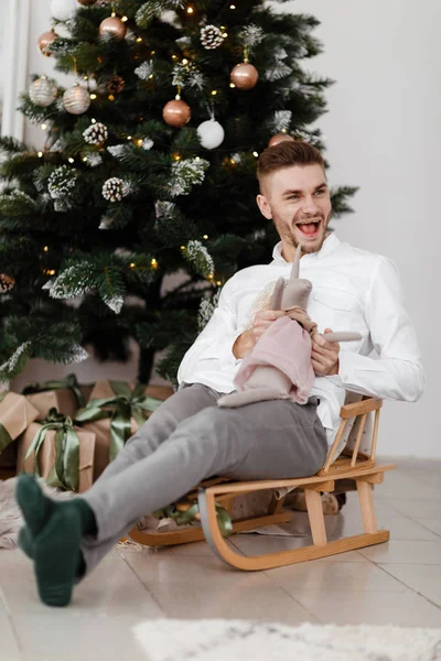 Homem alegre sentado no trenó em casa perto da árvore de Natal. um homem num trenó. Bom humor de Natal. Conceito de família e férias . — Fotografia de Stock