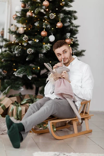 Uomo allegro seduto sulla slitta a casa vicino all'albero di Natale. uomo su una slitta. Buon umore natalizio. Concetto famiglia e vacanza . — Foto Stock