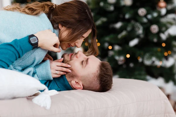 Casal apaixonado deitar-se na cama e quer beijar. guirlandas na parede e árvore de Natal perto da cama. O casal feliz está deitado na cama juntos. Desfrutando da companhia uns dos outros . — Fotografia de Stock