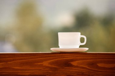 Tahta bir masada beyaz bir fincan kahve. Beyaz çay fincanı yeşil arka plandaki ahşap masada. Bahçe ve bir fincan kahve. Yazmak için yer.