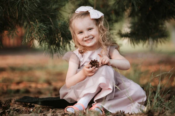 Pequena menina sorridente com um arco na cabeça e em um vestido com senta-se sob uma árvore. Criança gosta de brincar com cones — Fotografia de Stock