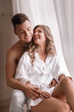 loving young couple enjoying morning at home near the window on Valentines Day. girl in white shirt and guy half naked having fun together